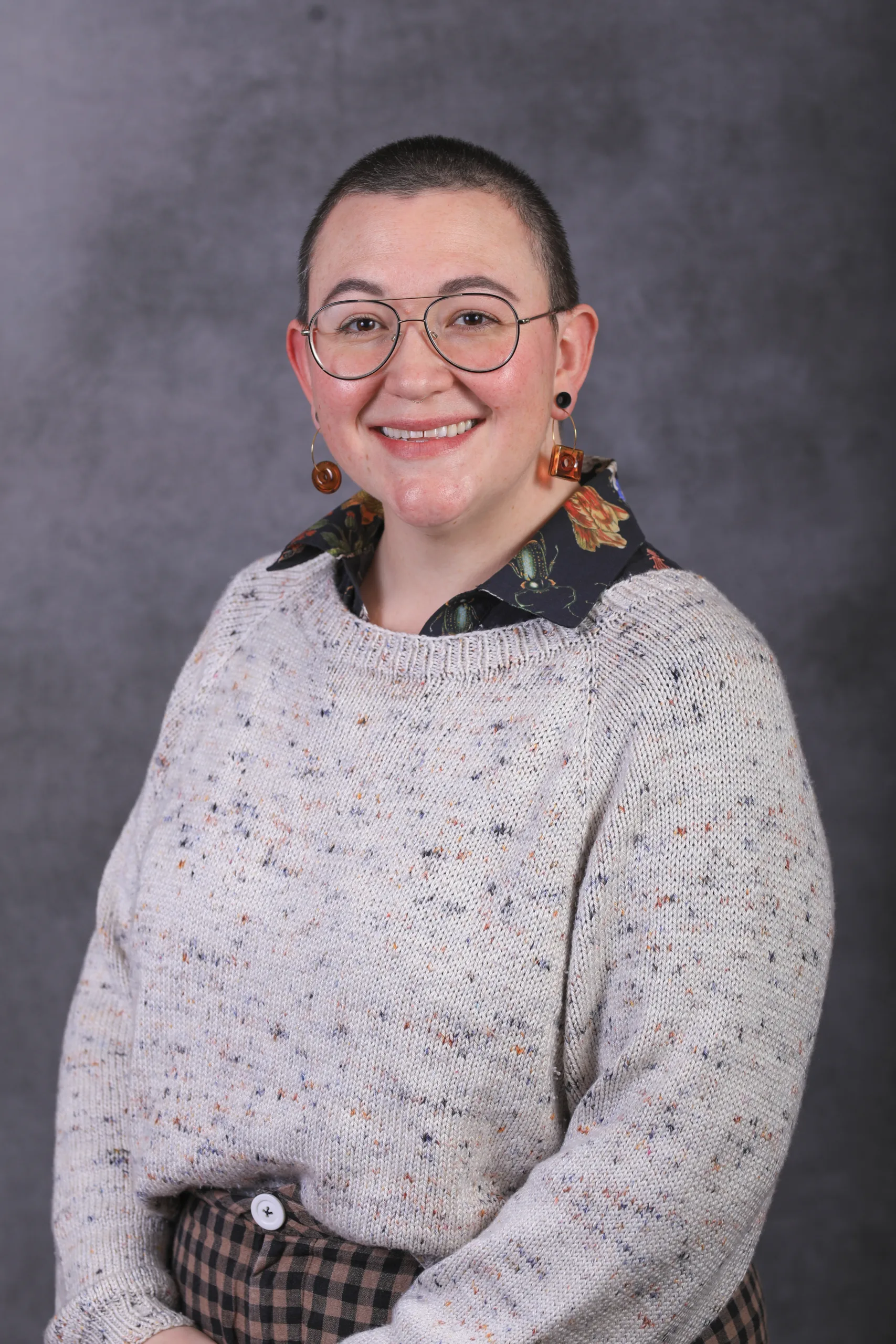 A person with short hair, glasses, and statement earrings smiles at the camera. They are wearing a light gray speckled sweater over a patterned shirt and plaid pants. The background is a gray gradient.
