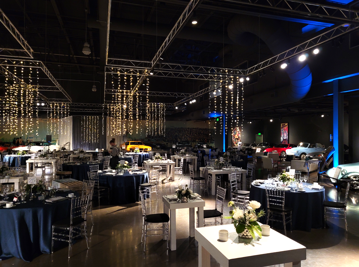 A spacious, dimly lit event hall with cars displayed along the walls. Round tables with blue tablecloths and silver chairs are set up for dining. String lights hang from the ceiling, creating a festive atmosphere. Sparse white floral centerpieces adorn the tables.