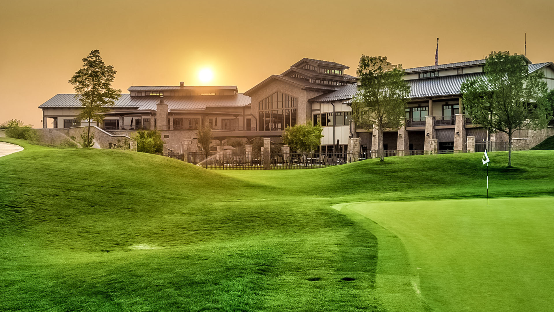 A golf course in the foreground with a well-maintained green is shown at sunset. Behind the course is a large, elegant clubhouse with tiered roofs and large windows. The sky is warm and orange as the sun sets behind the building. Trees are scattered around the scene.