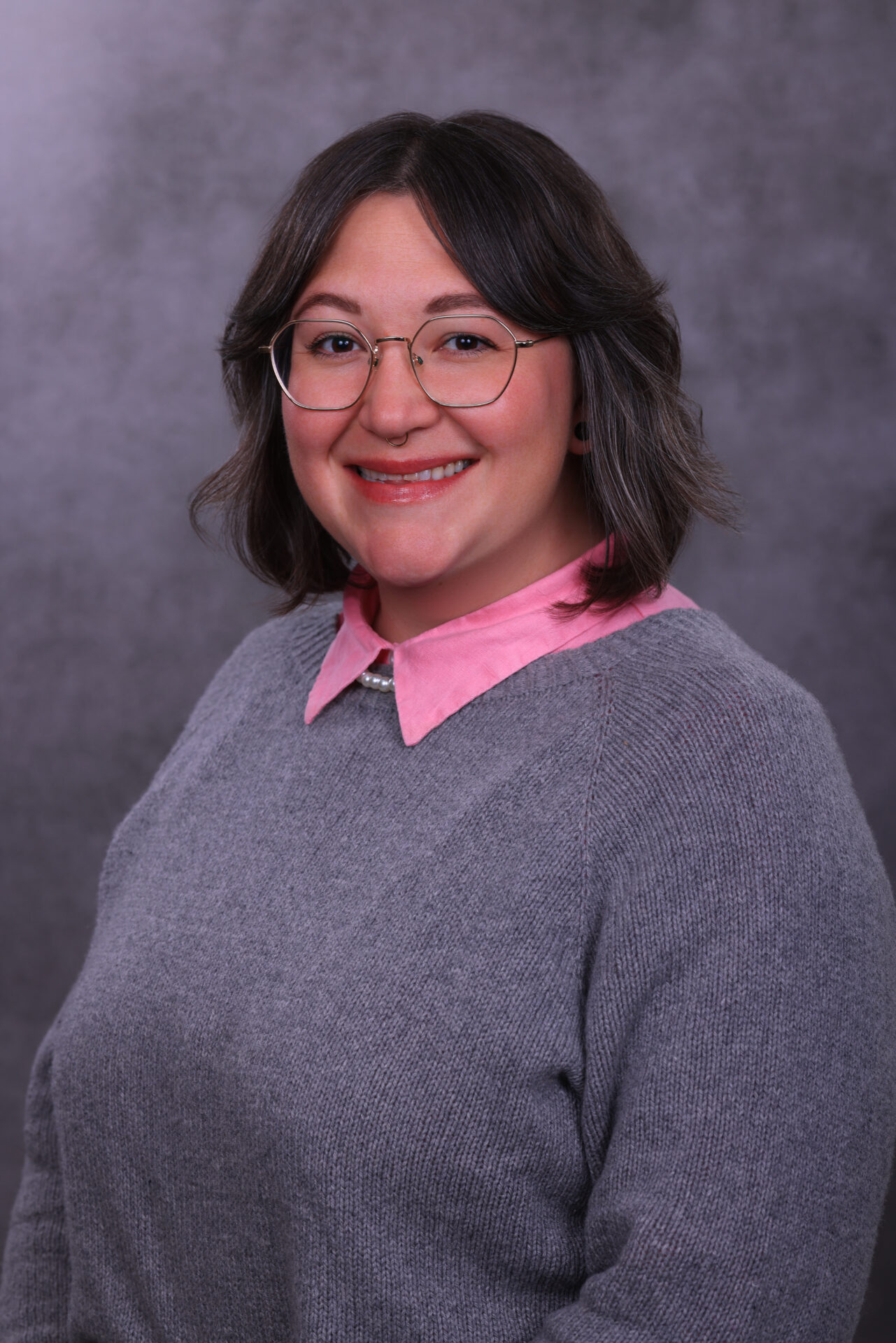 A person with short hair, glasses, and statement earrings smiles at the camera. They are wearing a light gray speckled sweater over a patterned shirt and plaid pants. The background is a gray gradient.